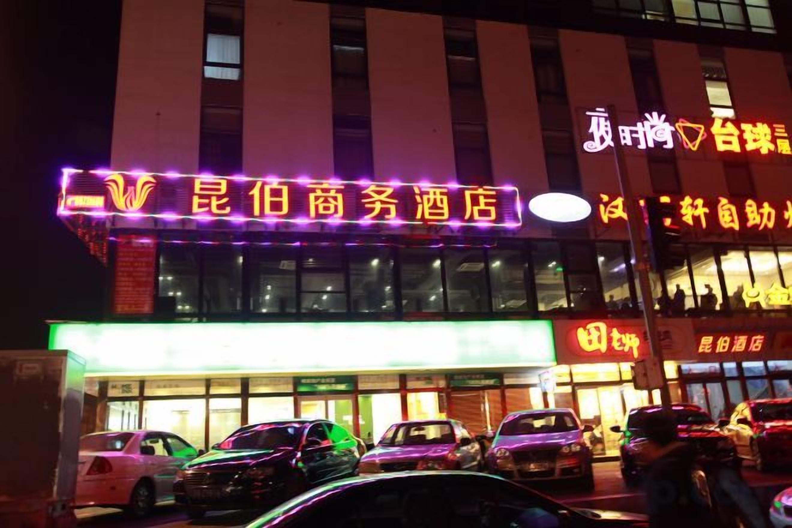 A hotel with a neon sign, multiple cars parked in front, and a building facade with windows.