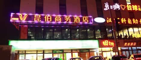 A hotel with a neon sign, multiple cars parked in front, and a building facade with windows.