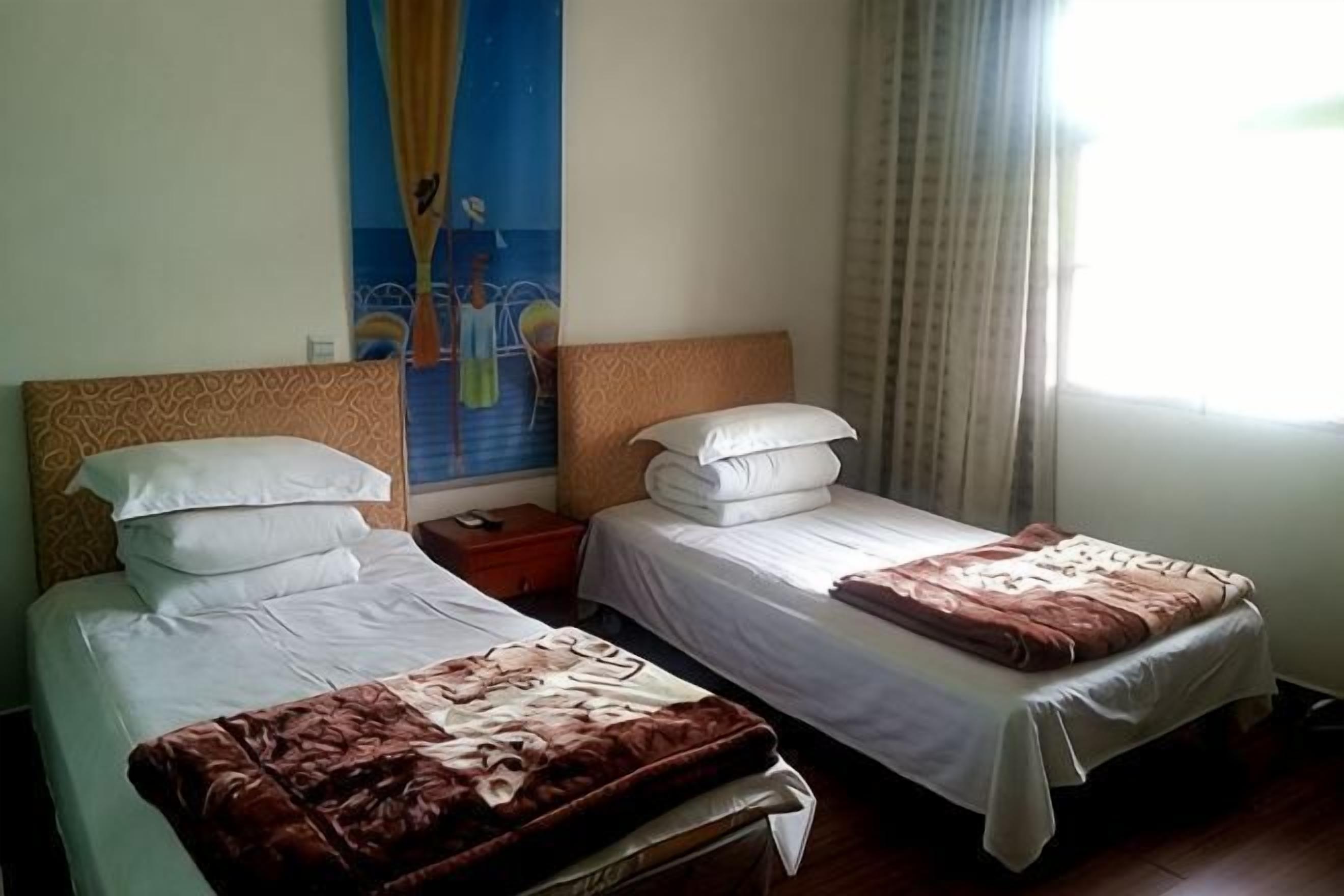 A hotel room with two beds, a nightstand, a painting on the wall, and a window with curtains.
