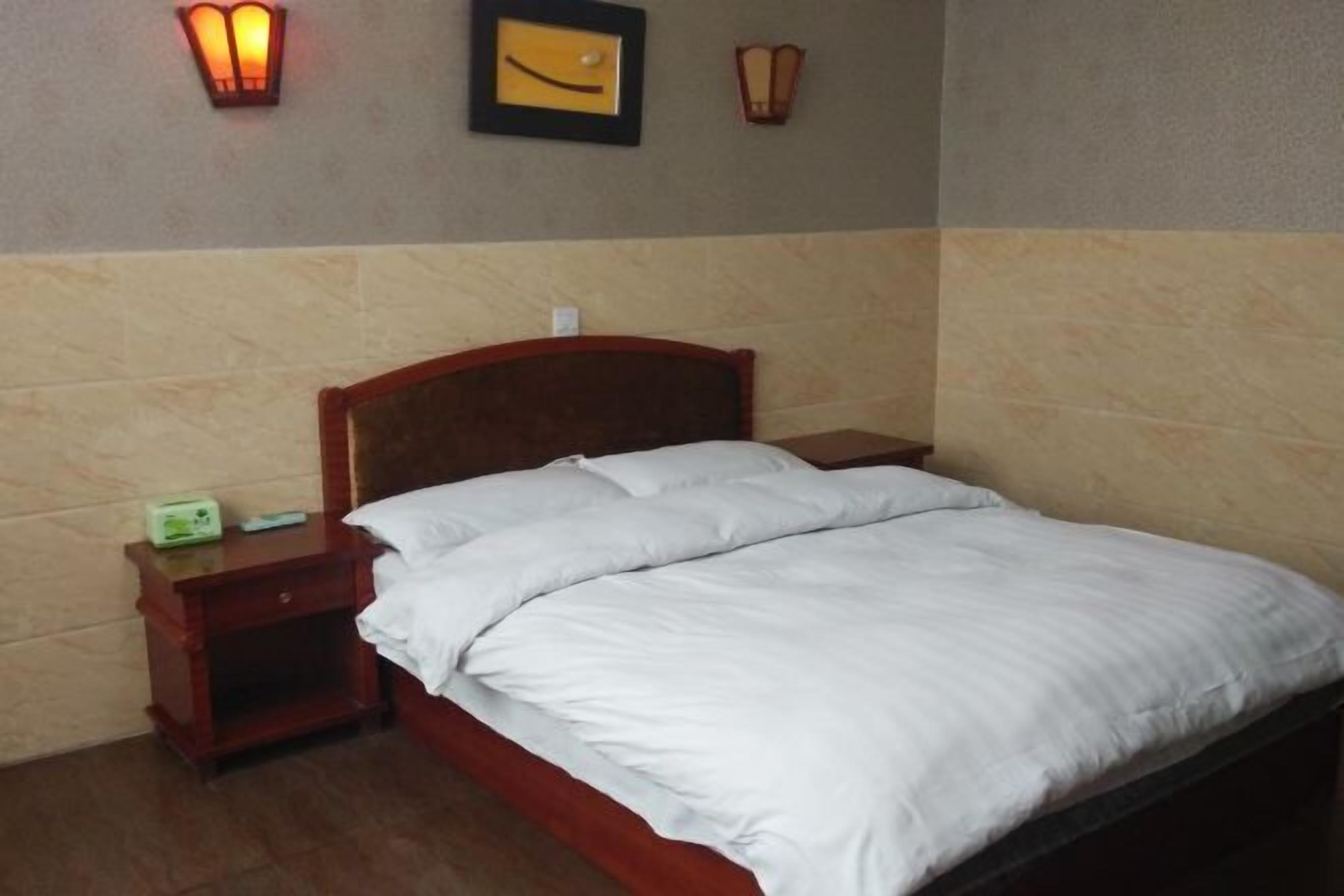 A hotel room with a wooden headboard, white bedding, a bedside table with a lamp, and a wall-mounted light fixture.