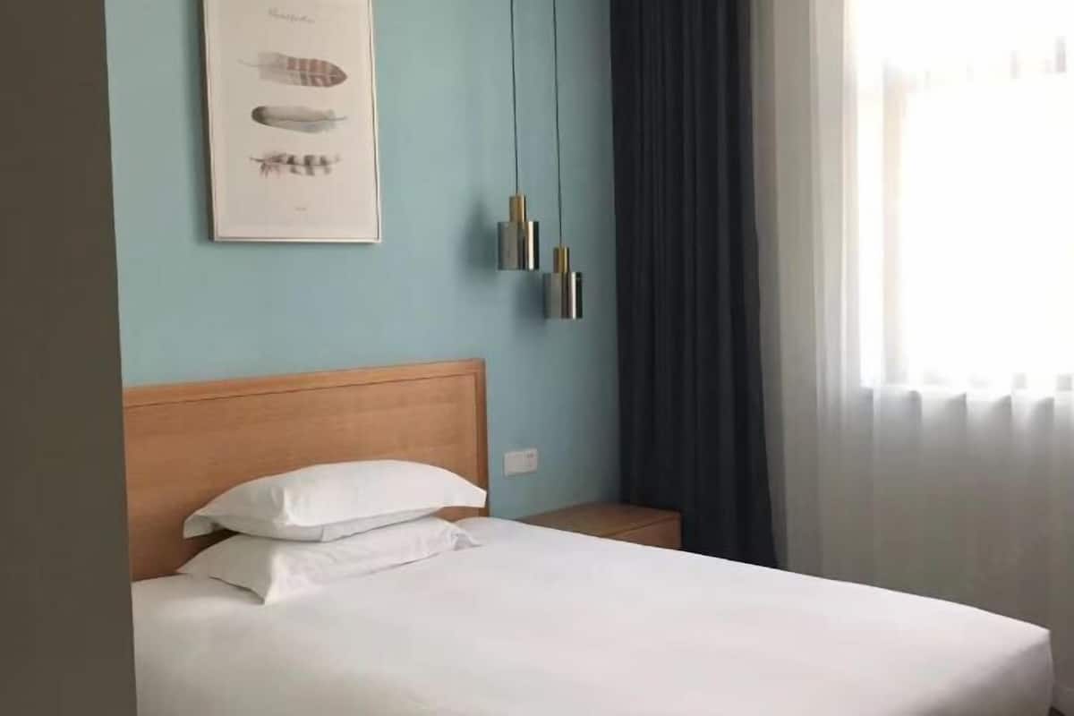 Un lit bien fait, avec du linge de lit blanc, une tête de lit en bois et un tableau représentant des plumes d’oiseaux accroché au mur.