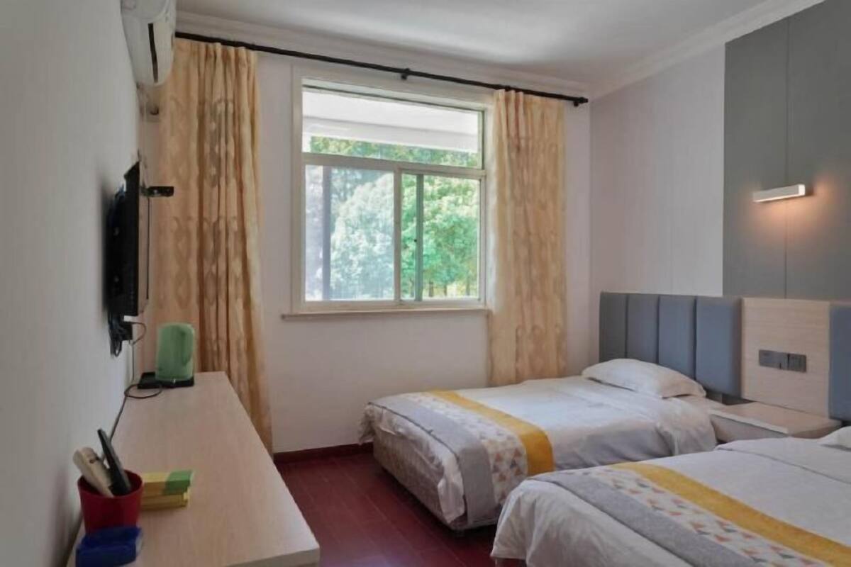 Une chambre avec deux lits, une fenêtre avec des rideaux, un bureau avec une lampe et un téléviseur fixé au mur.