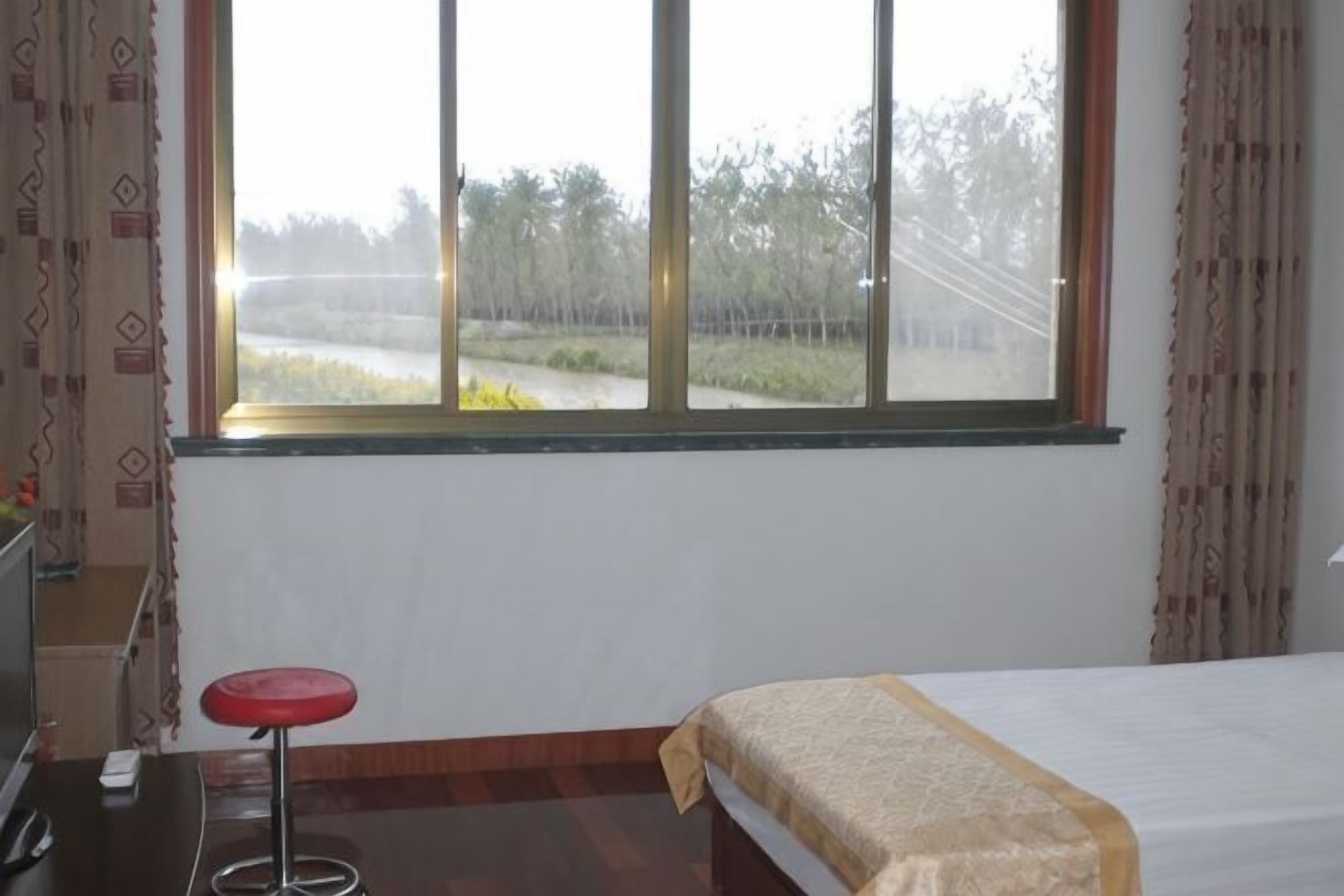 Una habitación con una cama, un taburete rojo, una ventana con vista a árboles y una cerca, y un escritorio con una silla.