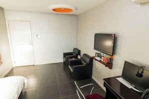 Quarto de hotel com cama, escrivaninha, televisão e área de estar com cadeiras.