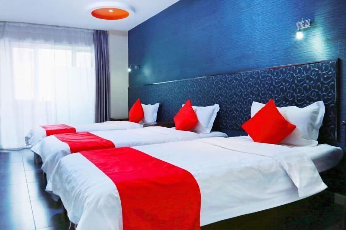 Habitación de hotel con dos camas, cada una con ropa de cama blanca y detalles rojos, una pared azul oscuro con textura e iluminación de techo moderna.