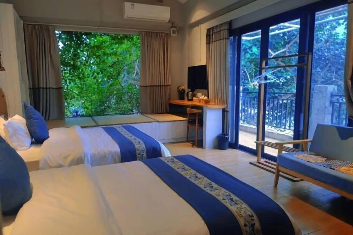 Une chambre d’hôtel avec deux lits, un bureau, une chaise et de grandes fenêtres donnant sur un paysage verdoyant.