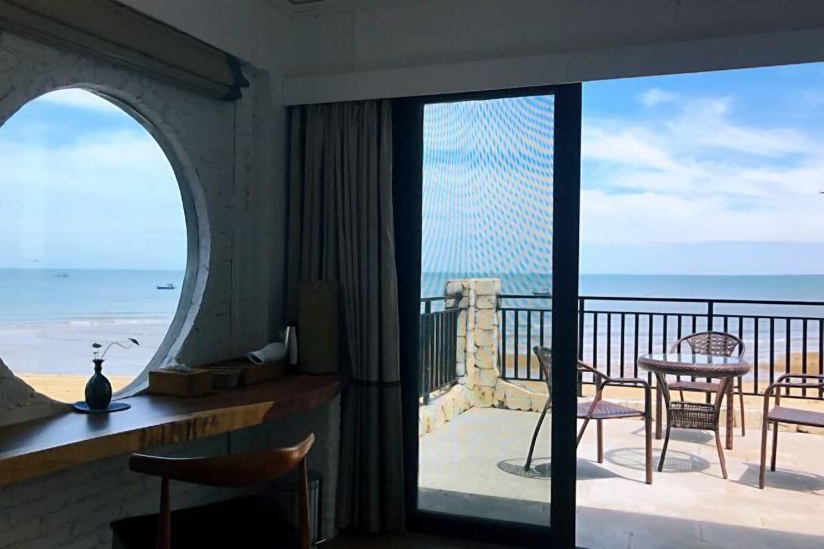 Una habitación con un ventanal grande, vista a la playa y un balcón con sillas y una mesa.