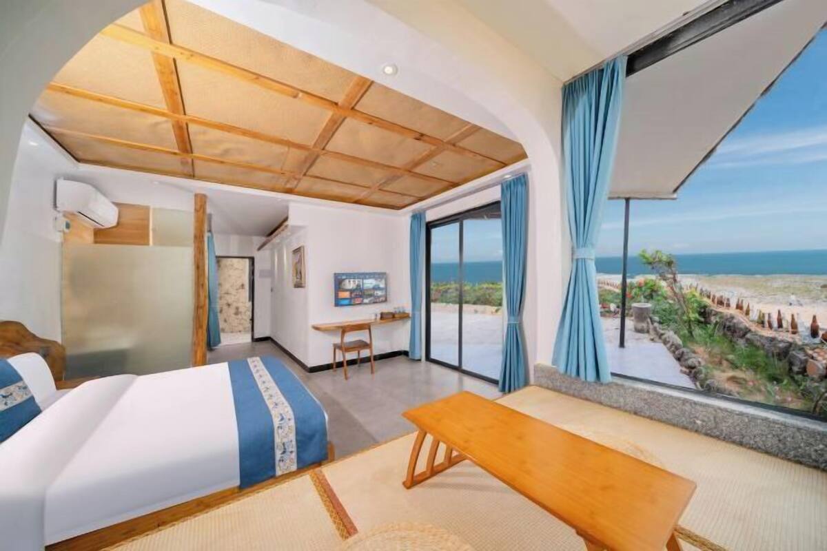 Une chambre d’hôtel avec un lit, une banquette, un bureau et une grande fenêtre donnant sur la plage.