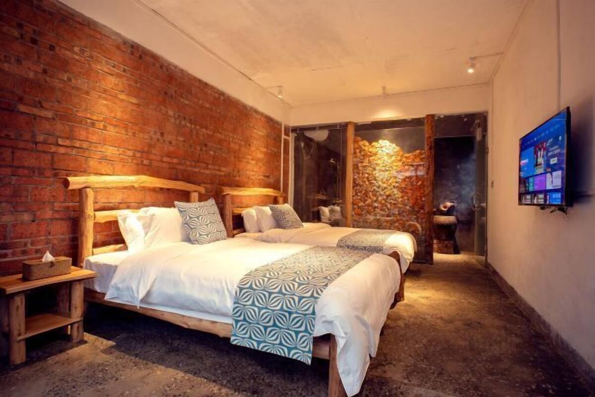 Une chambre avec deux lits, un mur en briques, une tête de lit en bois, une table de chevet et un téléviseur fixé au mur.
