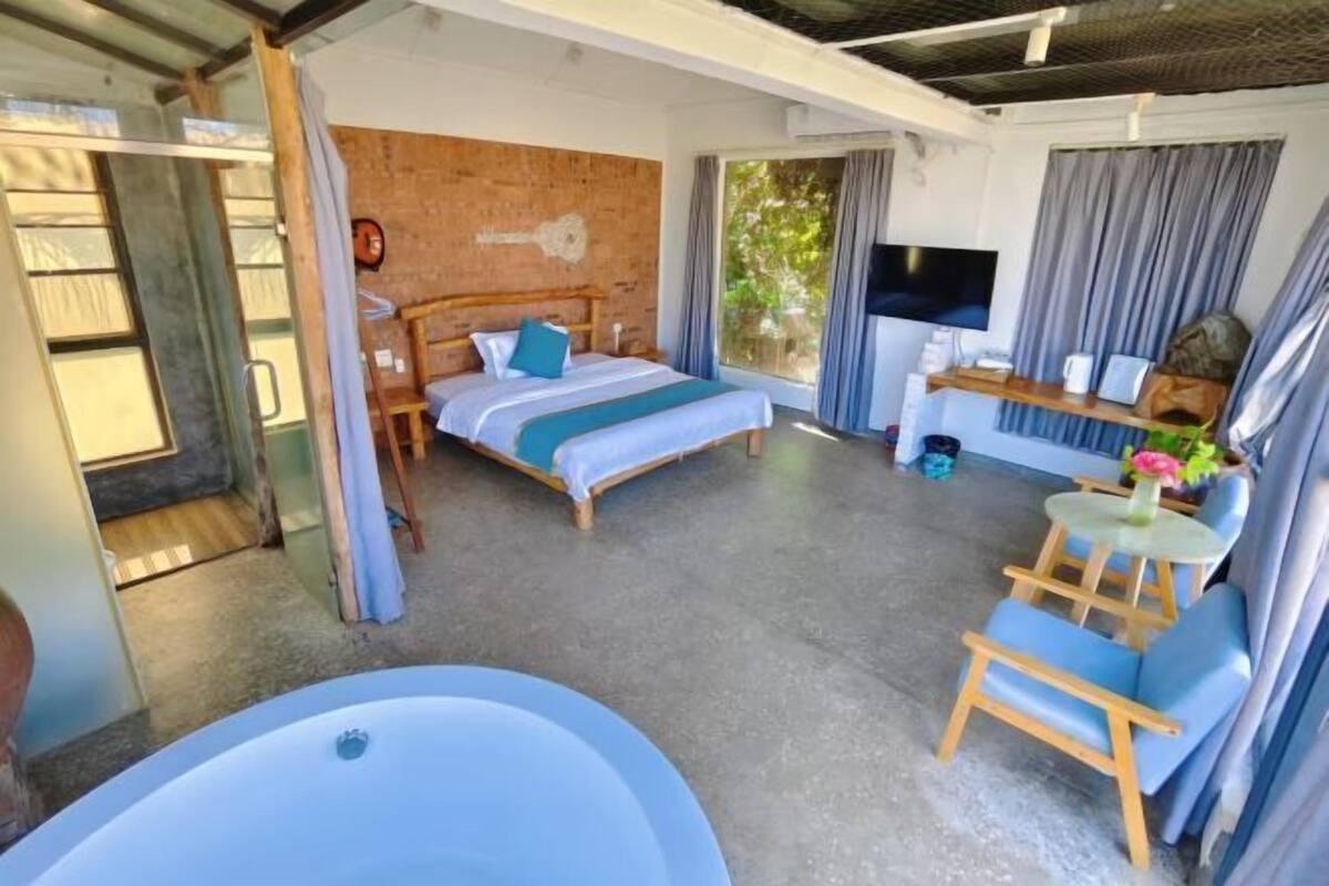 Une chambre à coucher avec un lit, une télévision, un bureau et une chaise. Il y a une fenêtre avec des rideaux et une vue sur les arbres.