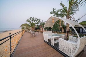 Una terraza al aire libre con muebles de mimbre blanco y una mesa de vidrio, con vista a la playa.