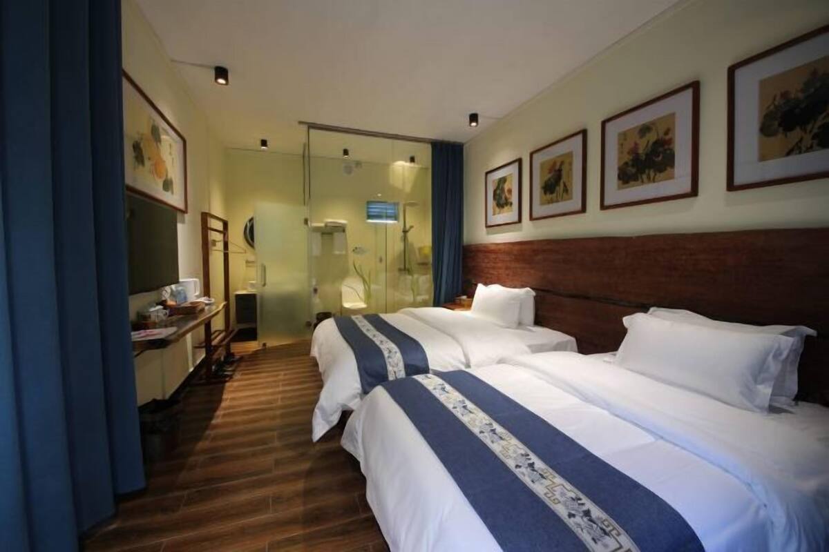 Une chambre d’hôtel avec deux lits, un plancher en bois, une salle de bain avec une porte en verre et des tableaux encadrés au mur.