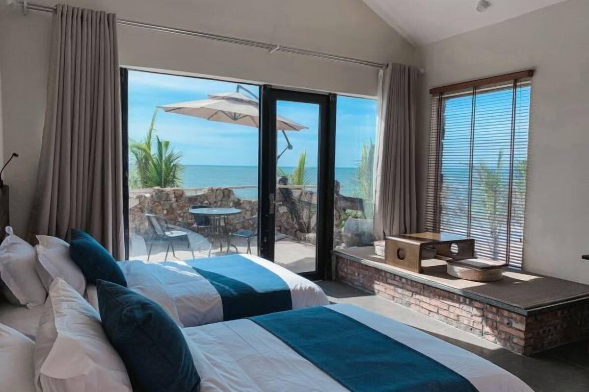 Une chambre avec deux lits, une vue sur l’océan et une terrasse avec des meubles d’extérieur.