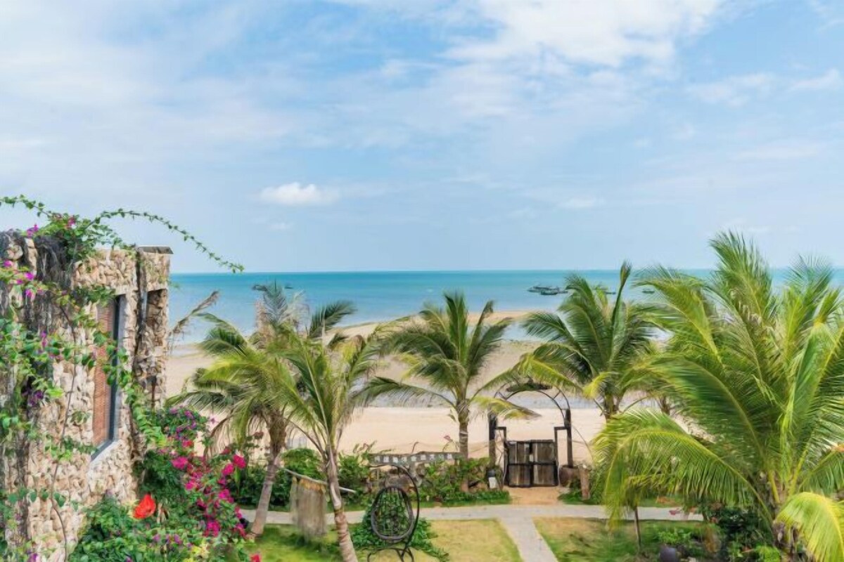 Vista de la playa con palmeras, un muro de piedra y un portón de madera.