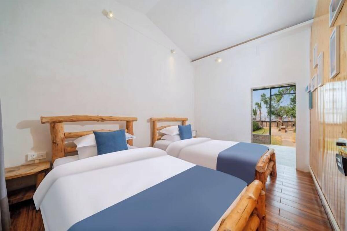 Une chambre avec deux lits, des têtes de lit en bois, des murs blancs, un plancher en bois et une vue sur l’extérieur.