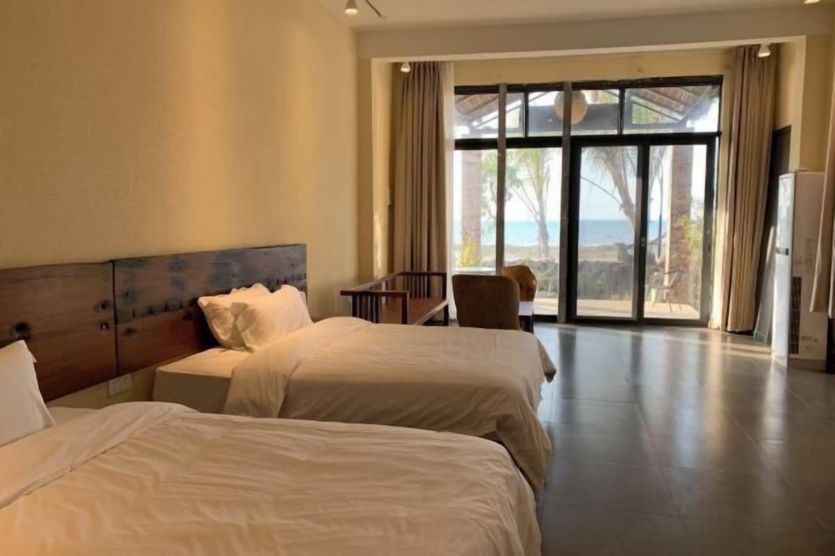 Quarto de hotel com duas camas, cabeceira de madeira, uma mesa pequena e uma grande porta de vidro deslizante que dá para uma varanda com vista para o oceano.