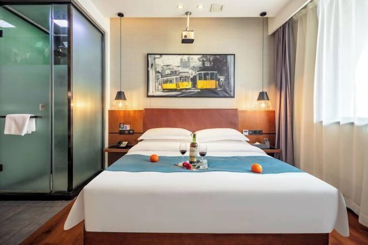 Een hotelkamer met een bed, een schilderij, twee nachtkastjes met lampen, een glazen douchecabine en een raam met gordijnen.
