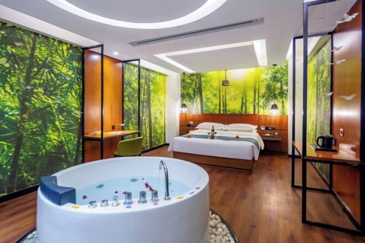 Een moderne hotelkamer met een groot bad, een bed, een bureau en uitzicht op een bos door de ramen.