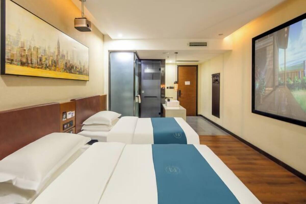 Een hotelkamer met twee bedden, een stadslandschap schilderij, een groot raam en een deur naar een badkamer.