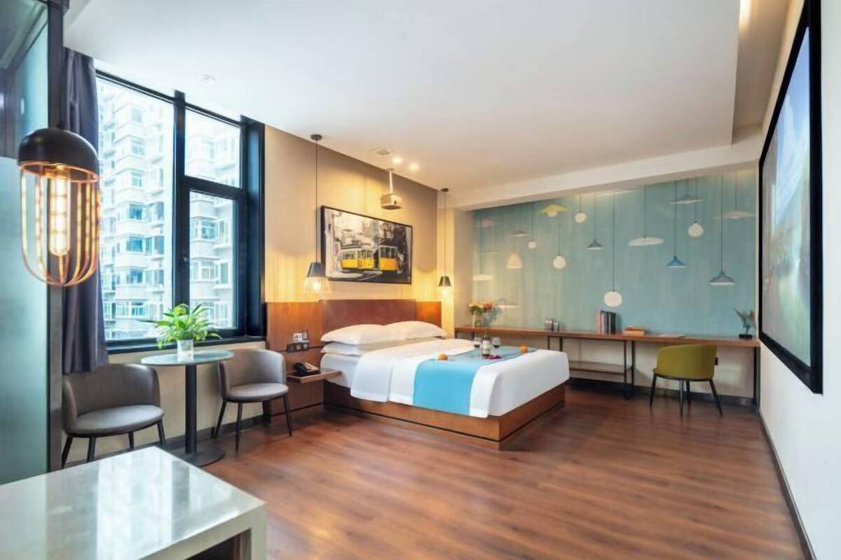 Een moderne hotelkamer met een bed, nachtkastje, stoelen, een bureau en een groot raam met uitzicht op gebouwen.