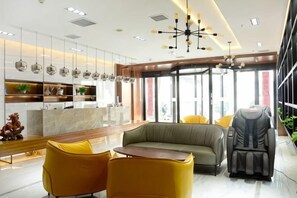 Un hall d’hôtel moderne comprenant un comptoir d’accueil, un espace salon, un fauteuil de massage et de grandes fenêtres.