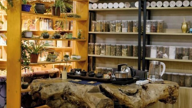 A cozy room with wooden shelves filled with plants and jars, a rustic wooden table with a kettle and teapot, and a shelf with neatly arranged cups and containers. - Dzongsa Book bar Inn (Shangri-La)