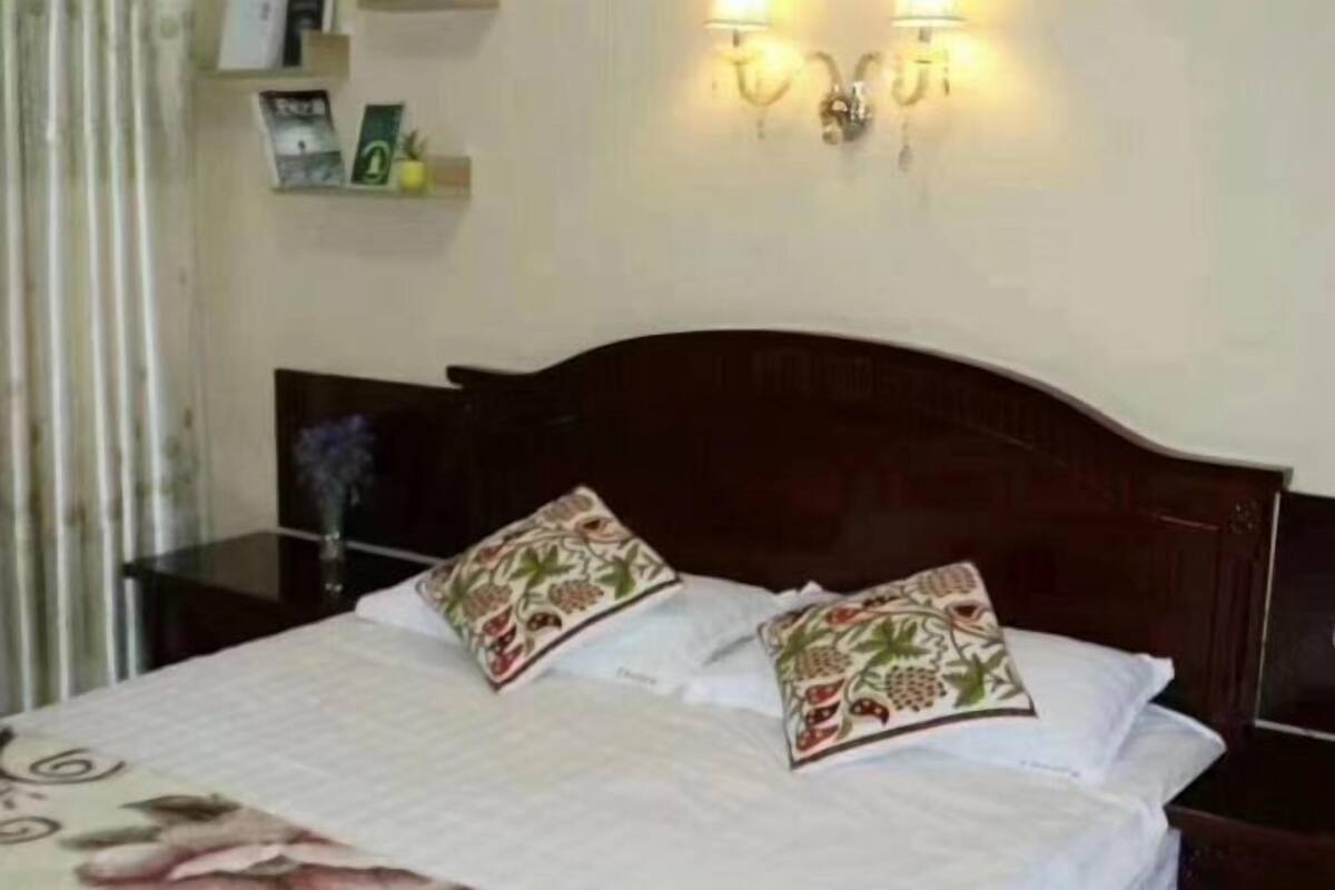 Una cama con colcha estampada, dos almohadones decorativos, una mesita de noche con un florero y apliques de pared.