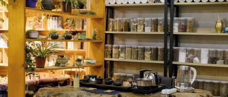 Un interior rústico con estantes de madera llenos de plantas y frascos, una mesa de madera con un hervidor, y una pared rústica con recipientes de almacenamiento.