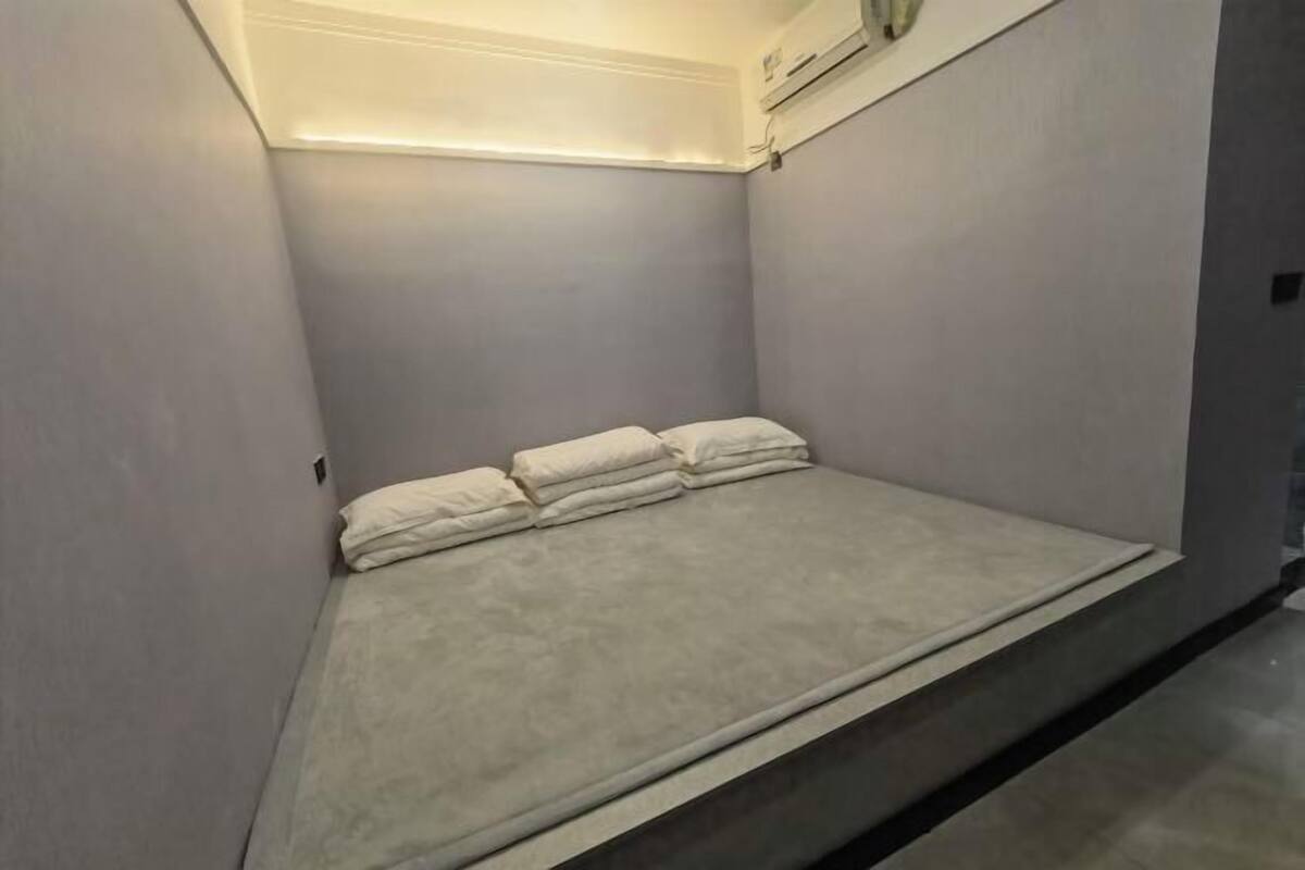 Una stanza piccola e scarsamente illuminata, con un letto singolo, un condizionatore d'aria a parete e un lampadario sopra il letto.