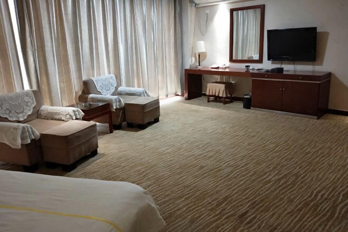 A hotel room with a large bed, two armchairs, a small sofa, a desk with a lamp, a television, and a large window with curtains.