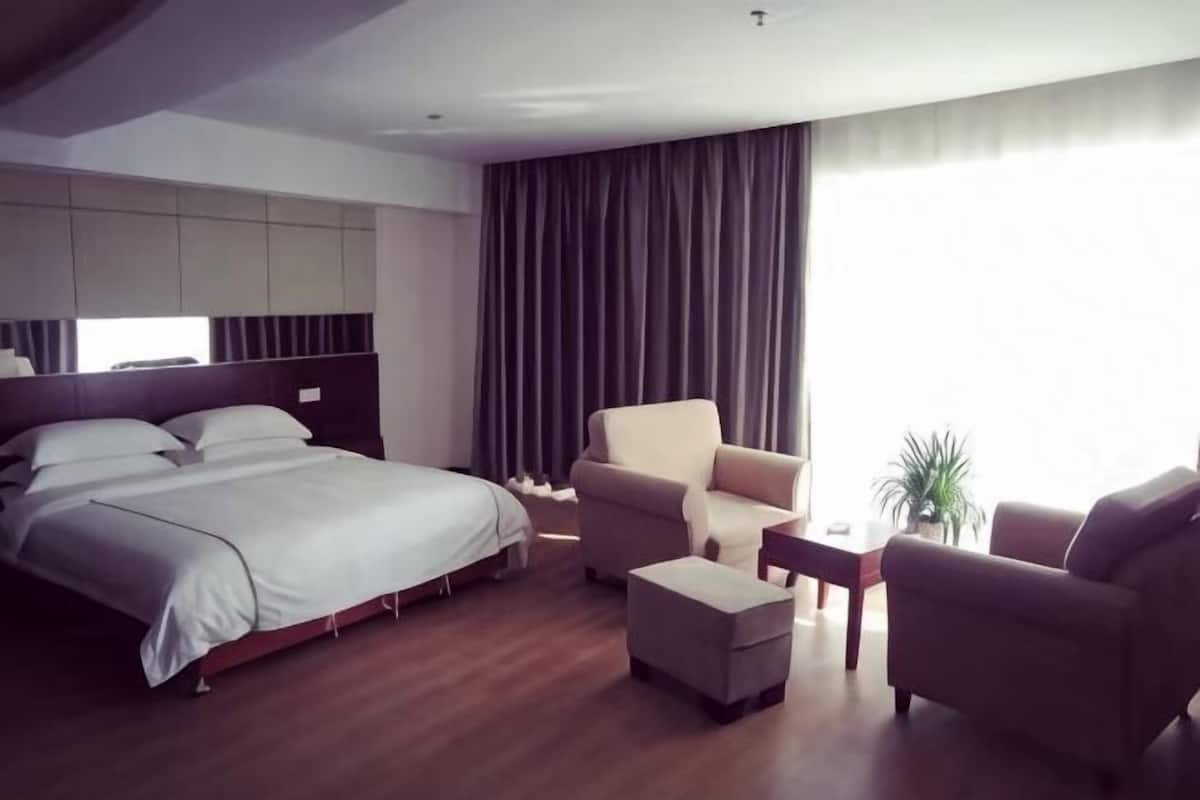 Una habitación de hotel moderna con una cama grande, dos sillones, una mesita y un ventanal con cortinas.