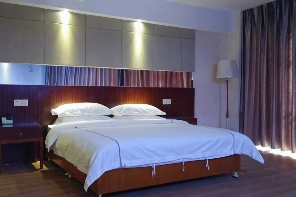 Habitación de hotel con una cama grande, una mesita de noche, una lámpara y vistas al exterior a través de cortinas.