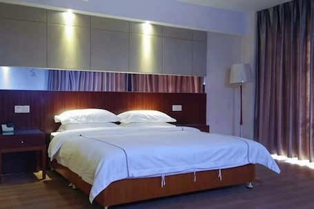 A hotel room with a large bed, a nightstand, a lamp, and a view of the outside through curtains.. Dihao Hotel