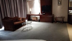 Une chambre d’hôtel comprenant un canapé, une table basse en verre, un bureau avec une chaise, une télévision et une petite table avec une lampe.