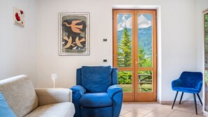A living room with a sofa, a blue armchair, a blue chair, a door leading to a balcony with a mountain view, and two framed artworks on the wall.