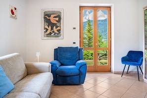 A living room with a sofa, a blue armchair, a blue chair, a door leading to a balcony with a mountain view, and two framed artworks on the wall.