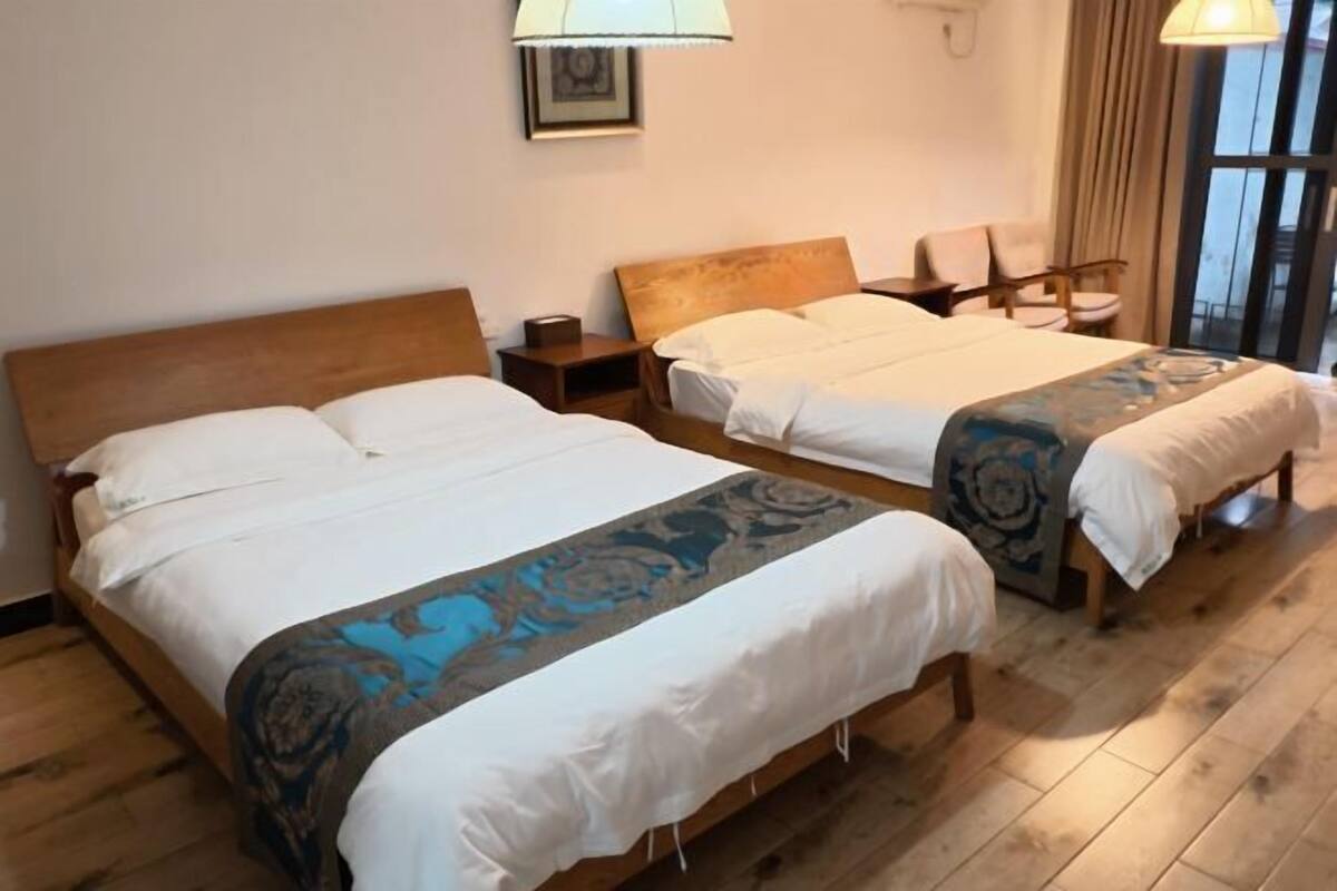 Una habitación de hotel con dos camas, cabeceros de madera, mesitas de noche, una silla, una lámpara y una ventana con cortinas.