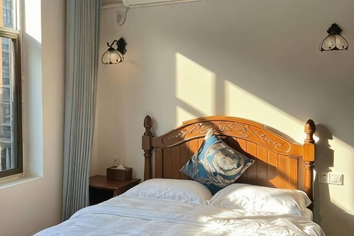 Un dormitorio con cabecera de madera, una cama individual con ropa de cama blanca, una mesita de noche con lámpara y una ventana con cortinas.