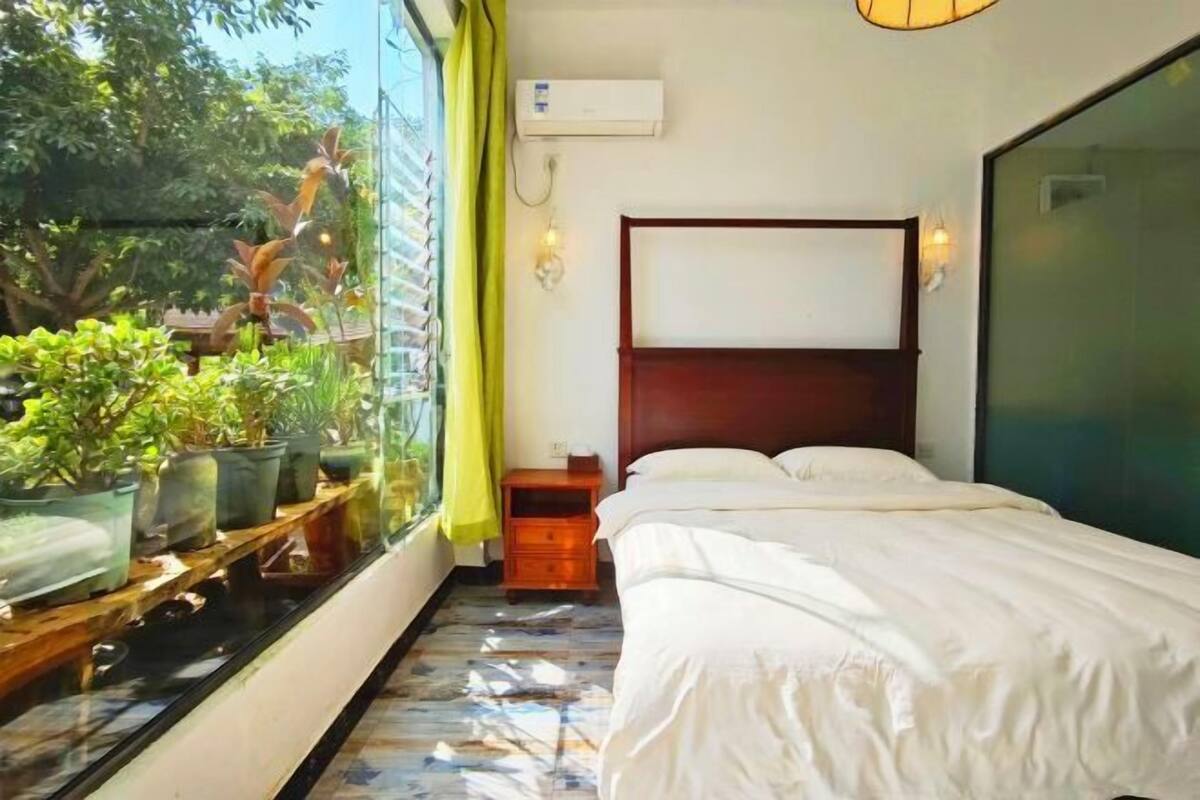 Un dormitorio con cama, mesita de noche, ventana con plantas y equipo de aire acondicionado.