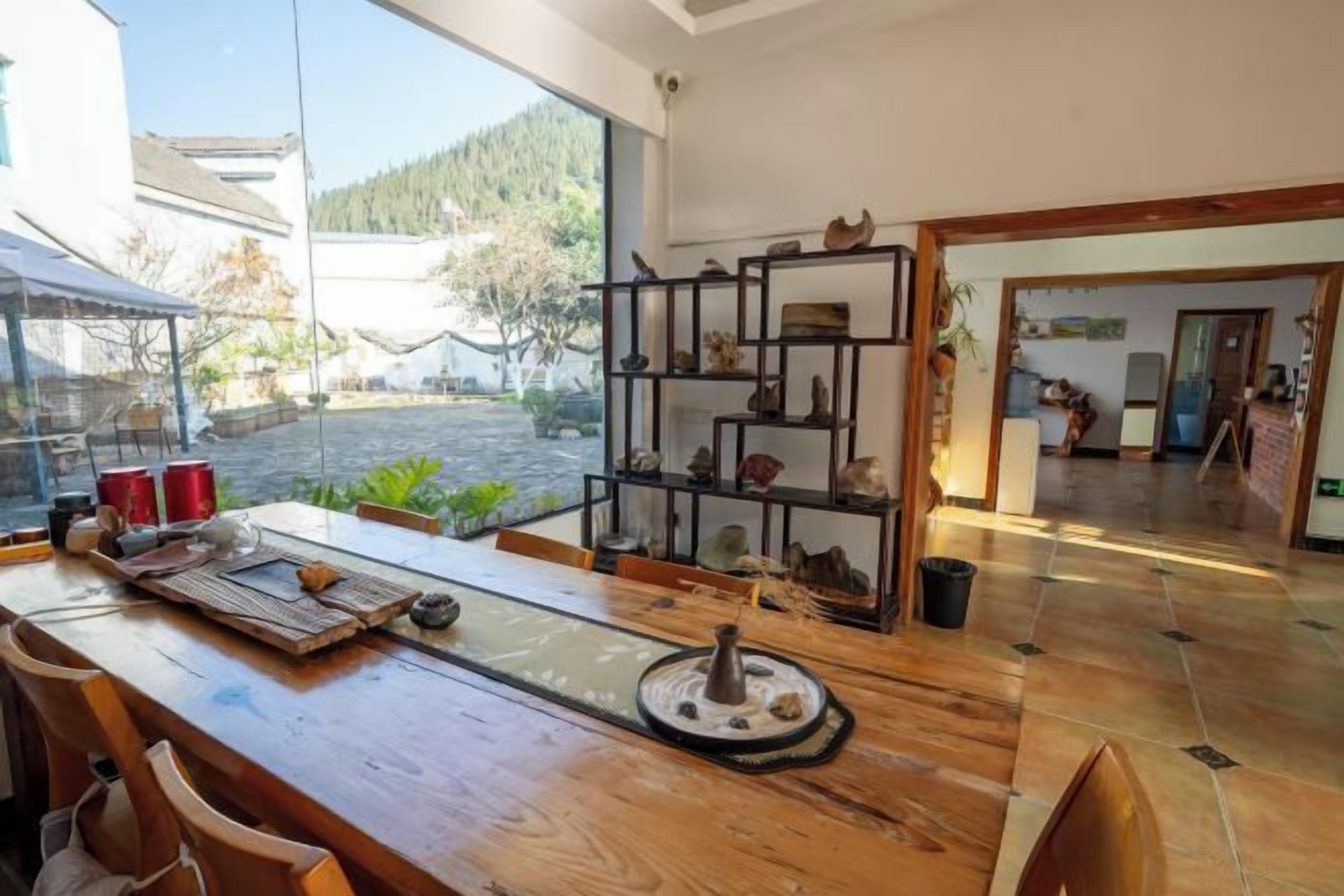 Una mesa de comedor de madera con sillas, un estante con adornos, y vistas a un jardín y un edificio.