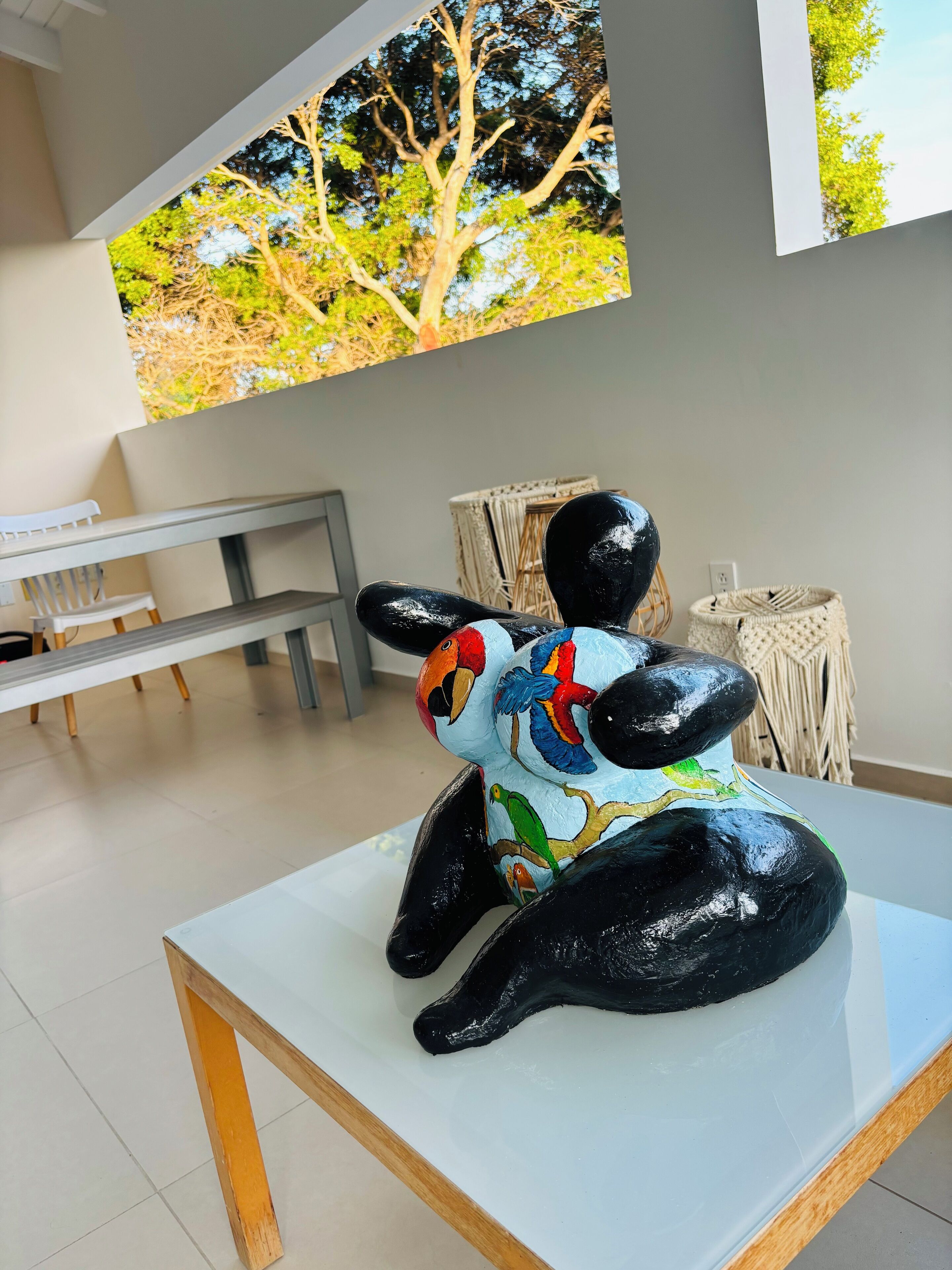 A sculpture of a bird with colorful feathers on a white table in a room with a bench and a woven basket.