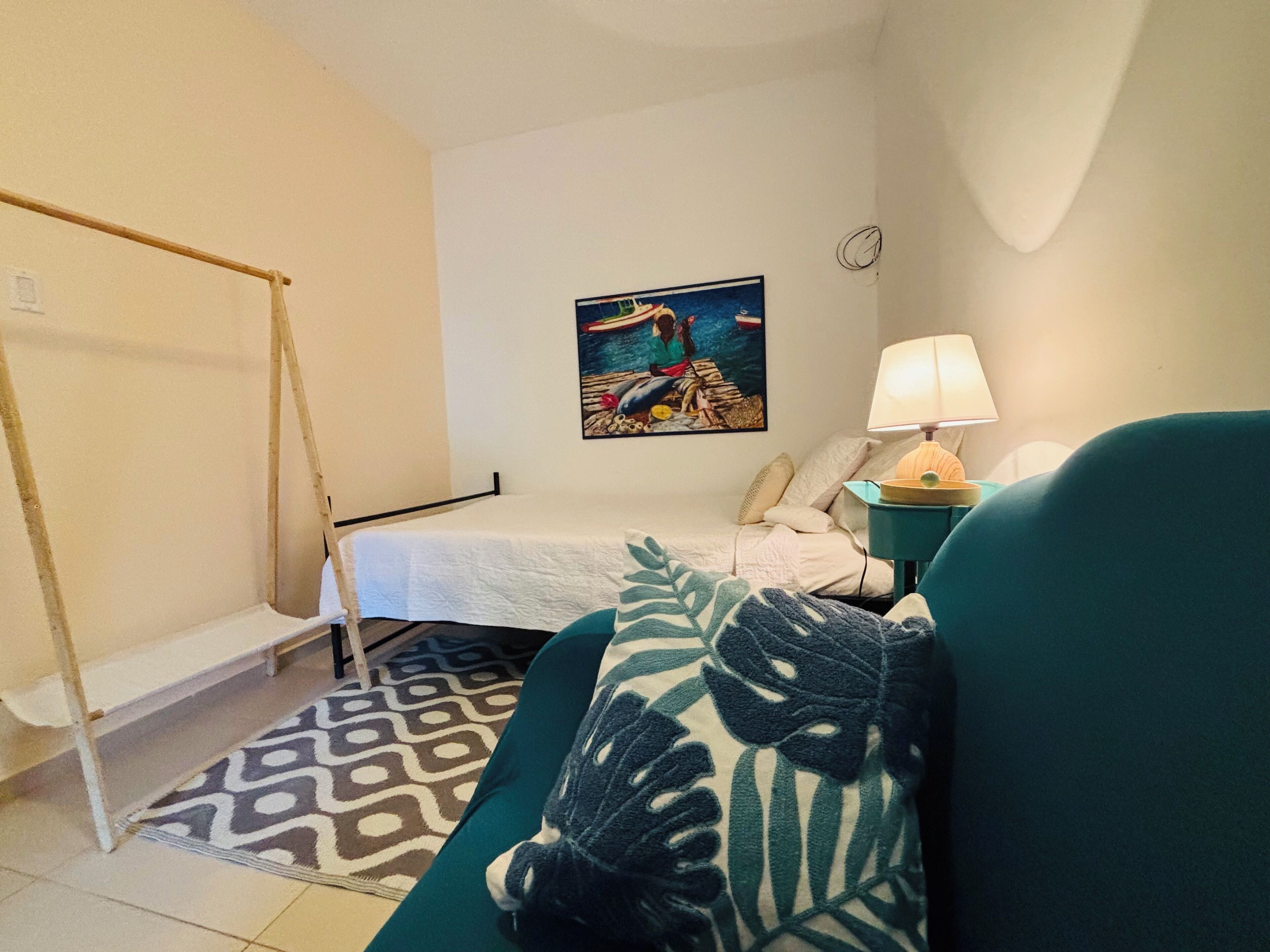 A bedroom with a bed, a green sofa, a patterned rug, a painting on the wall, and a lamp on a side table.