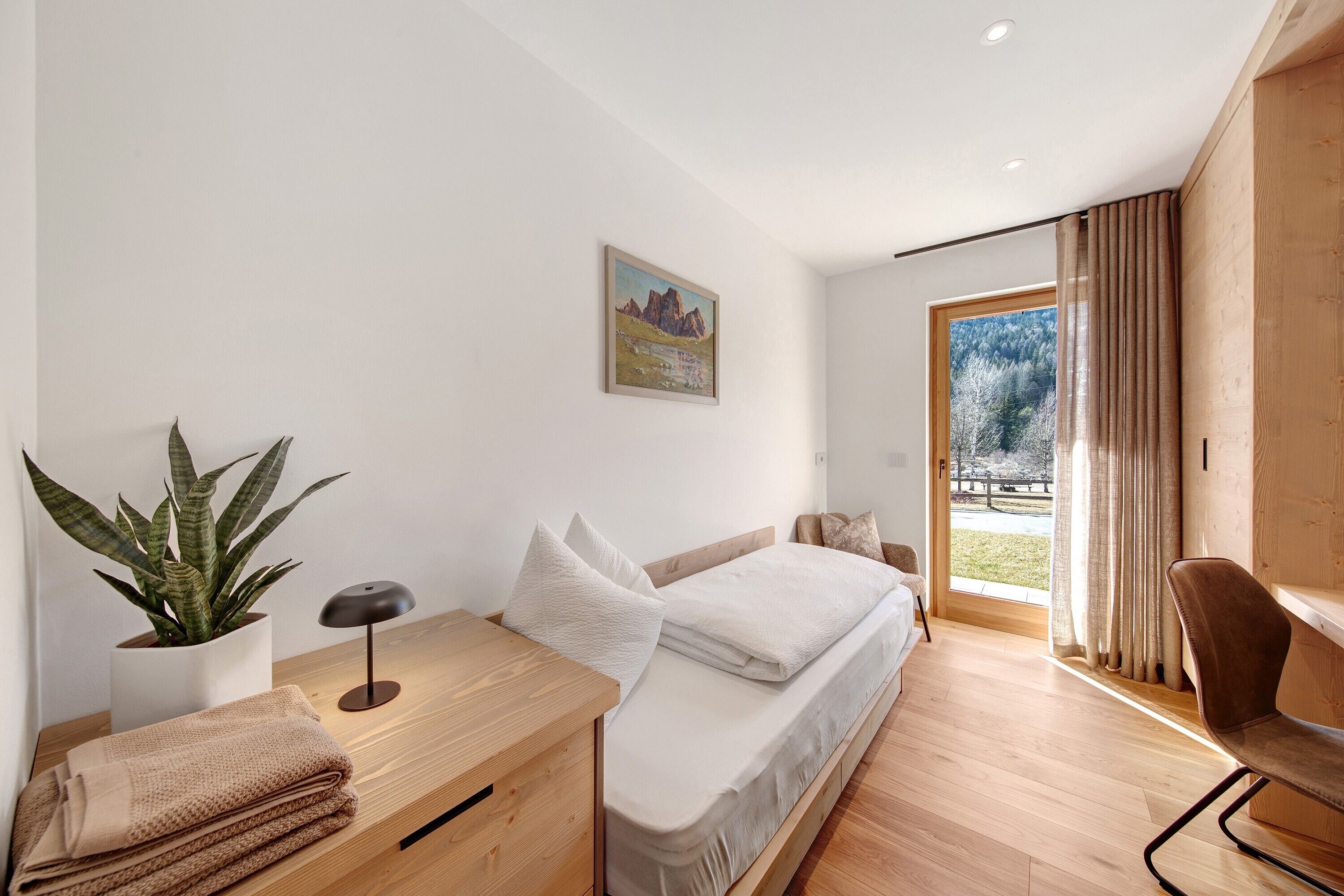 A modern bedroom with a bed, bedside table, lamp, potted plant, and a view of the outdoors.
