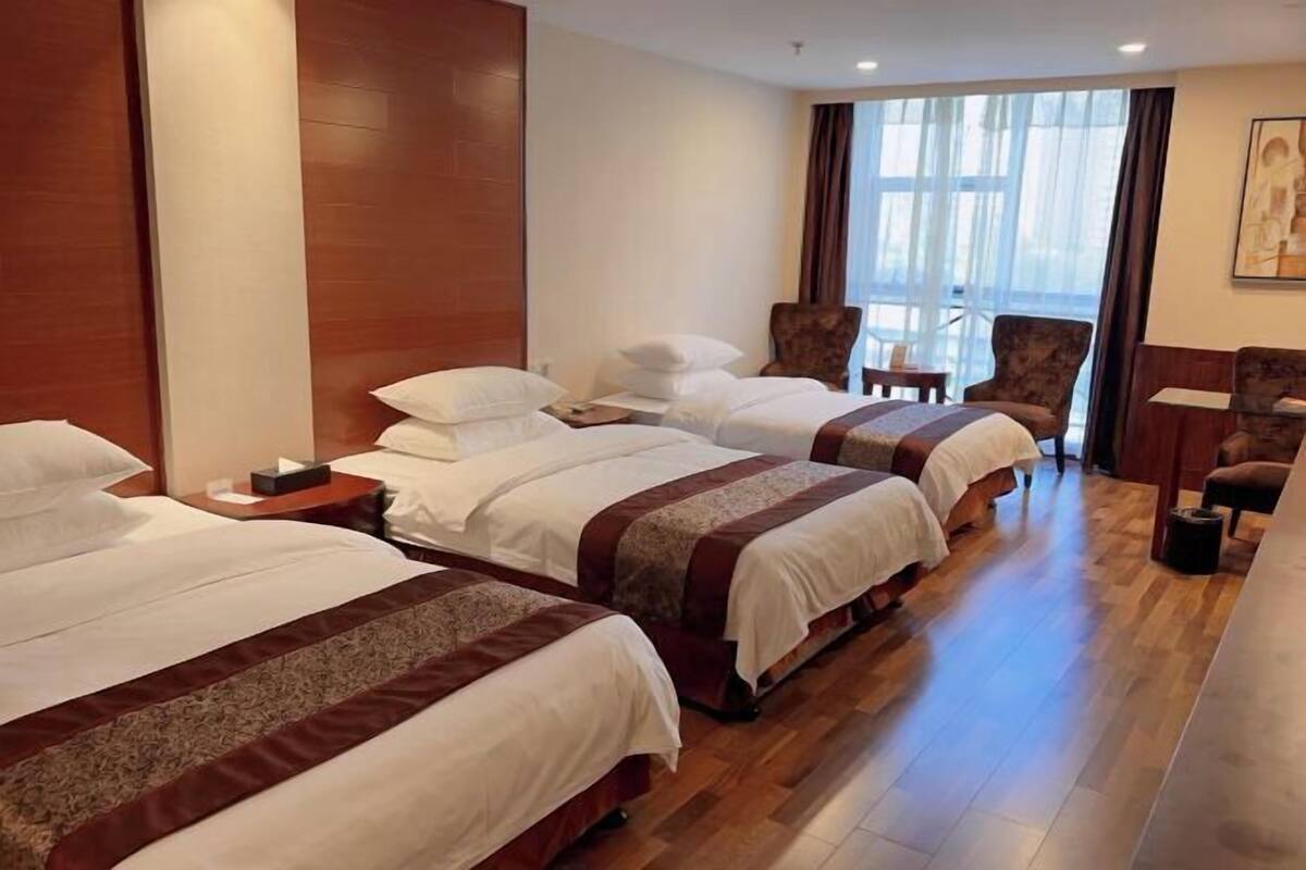 A hotel room with three beds, wooden flooring, a large window with curtains, and a seating area with chairs.
