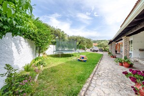 Un chemin pavé mène à une maison dont le mur est blanc et qui est bordée d’une pelouse verte.