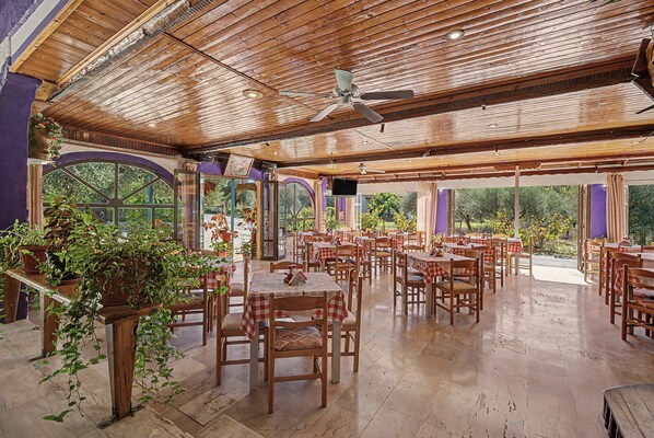 An indoor dining area with wooden tables and chairs, large windows, and a ceiling fan. - Studio Apartment 'Maltas  20' with Shared Terrace, Wi-Fi and Air Conditioning (Corfu)