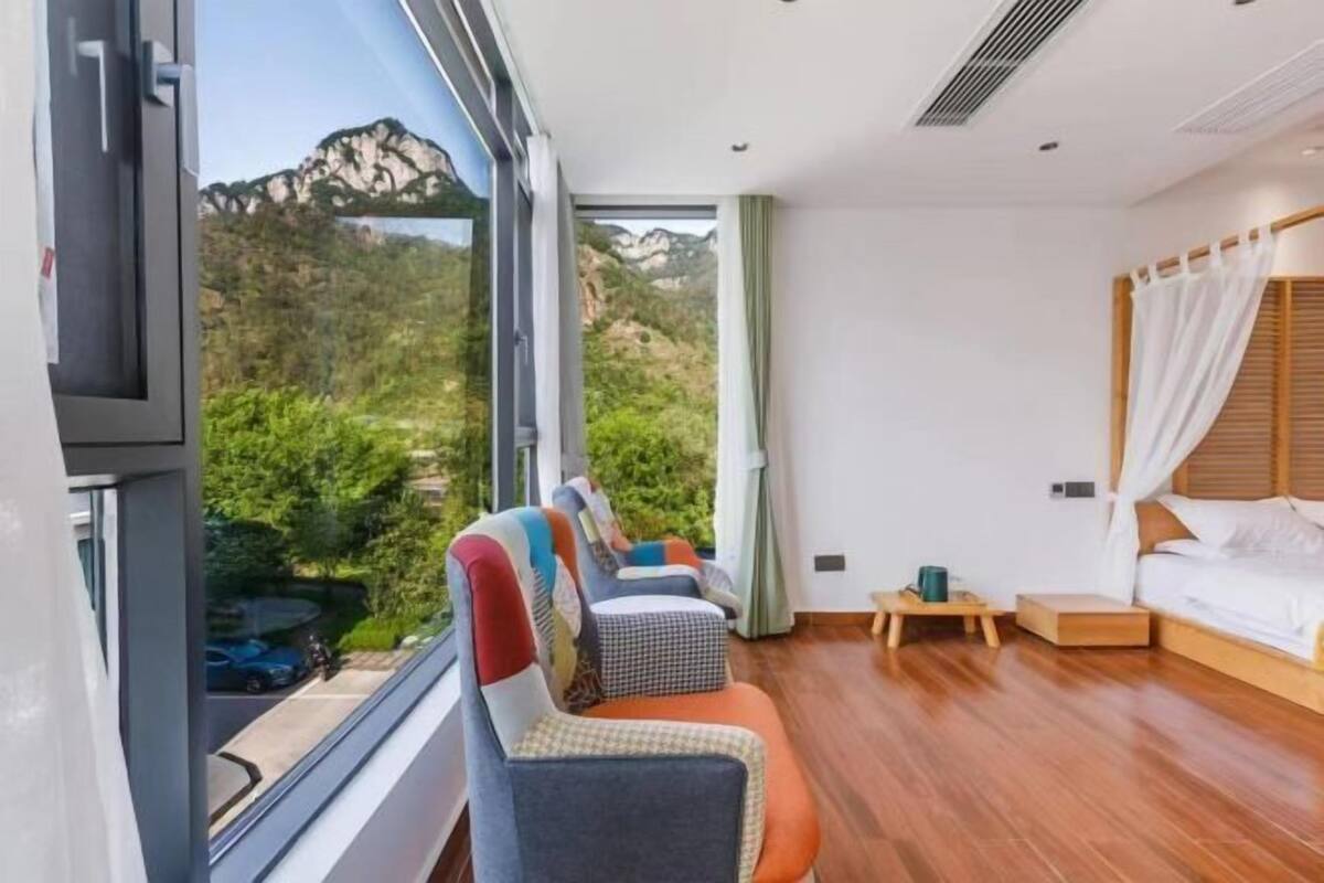 A modern hotel room with a large window, a bed, a bedside table, and a seating area with colorful chairs.