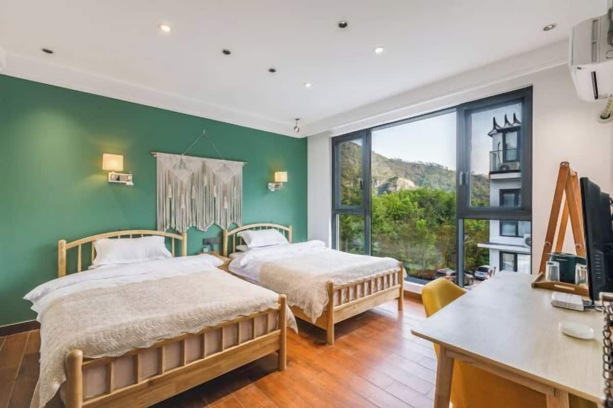 A room with two wooden beds, a desk, and a large window offering a view of greenery and buildings.