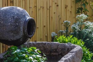 A large, rustic, black pot pouring water into a stone basin surrounded by lush greenery and a bamboo fence.