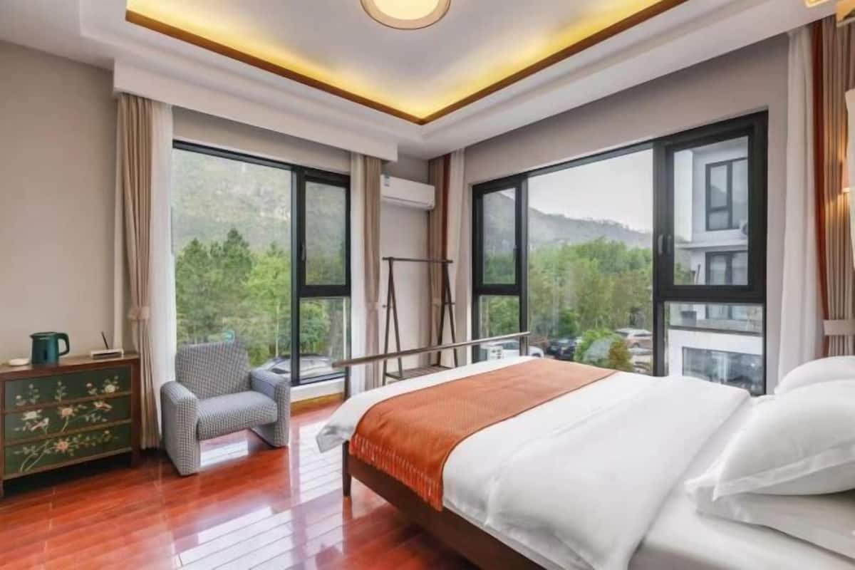 A bedroom with a bed, a chair, a dresser, and large windows with a view of greenery.